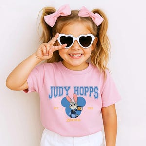 Op de afbeelding: Roze T-shirt met de blauwe tekst "JUDY HOPPS" en een cartoonafbeelding van Judy Hopps uit Zootopia. Het shirt heeft ook de tekst "EST. 2016" en "Zootopia". Het meisje draagt hartvormige zonnebrillen en roze strikken.