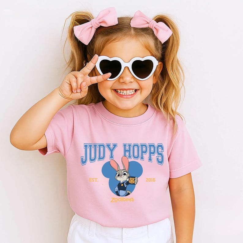 Op de afbeelding: Roze T-shirt met de blauwe tekst "JUDY HOPPS" en een cartoonafbeelding van Judy Hopps uit Zootopia. Het shirt heeft ook de tekst "EST. 2016" en "Zootopia". Het meisje draagt hartvormige zonnebrillen en roze strikken.