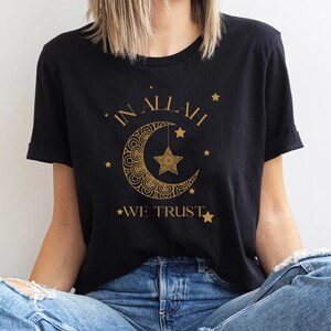 Może przedstawiać: Czarny T-shirt z nadrukiem złotego p&oacute;łksiężyca i gwiazdy. Na koszulce widnieje napis "IN ALLAH WE TRUST".
