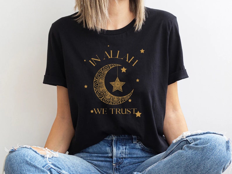 Może przedstawiać: Czarny T-shirt z nadrukiem złotego p&oacute;łksiężyca i gwiazdy. Na koszulce widnieje napis "IN ALLAH WE TRUST".