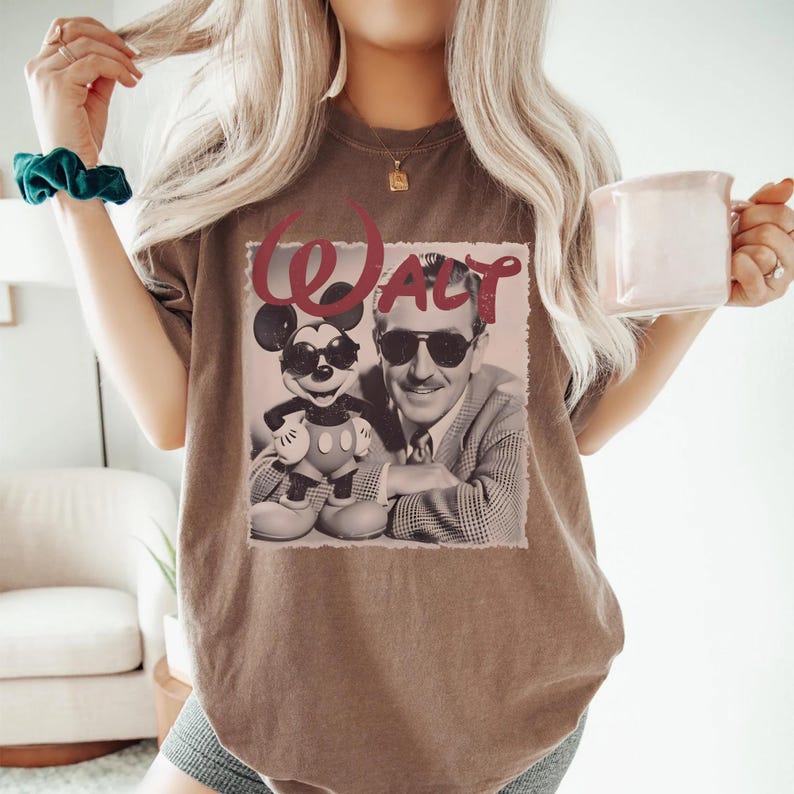 May include: A brown short-sleeve t-shirt featuring a graphic print of a man in a suit and a cartoon mouse, both wearing sunglasses. The word "WALT" is printed in large red letters above the figures, set against a light-colored rectangular background.