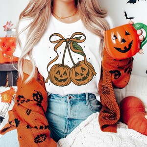 May include: White t-shirt featuring a Halloween-themed design of two pumpkin-shaped cherries with smiling faces, tied together with a brown ribbon. The person is wearing an orange sweater and holding an orange pumpkin mug.