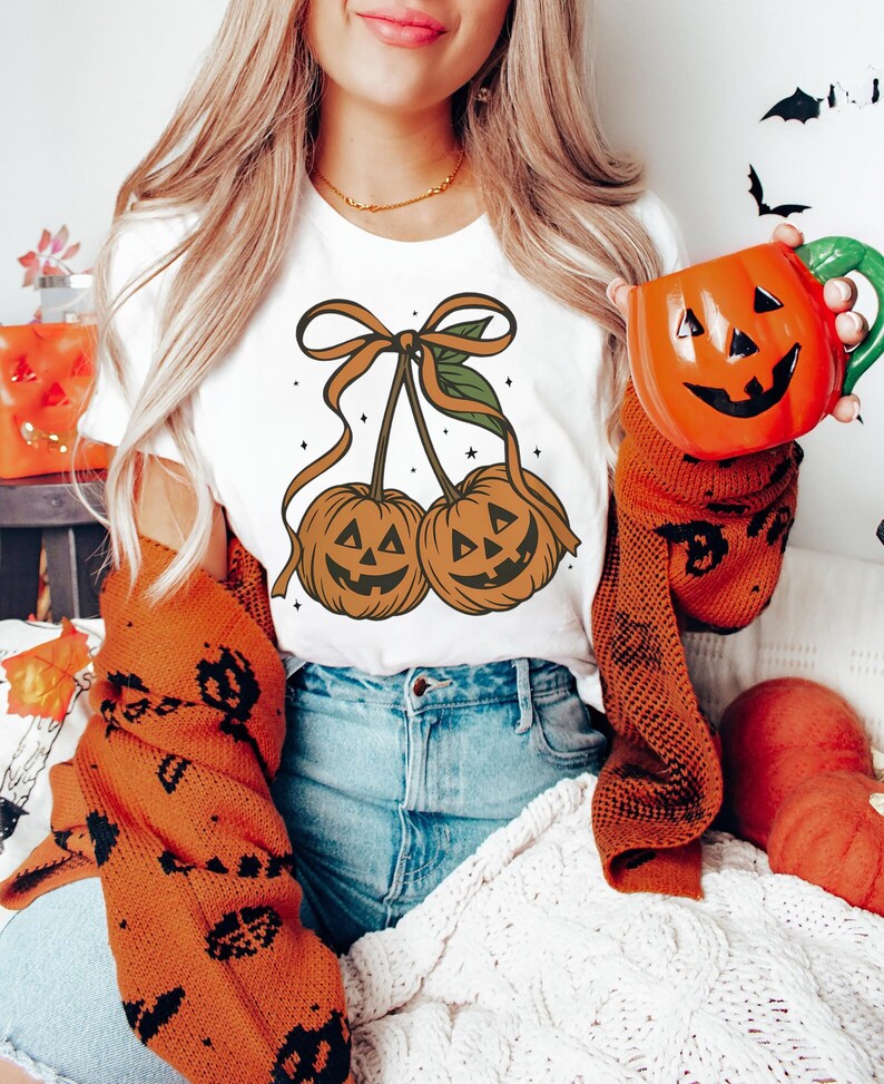 May include: White t-shirt featuring a Halloween-themed design of two pumpkin-shaped cherries with smiling faces, tied together with a brown ribbon. The person is wearing an orange sweater and holding an orange pumpkin mug.