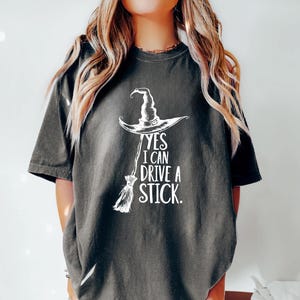 以下が含まれることがあります： 白い魔女の帽子とほうきのグラフィックと「YES I CAN DRIVE A STICK」のテキストが特徴の濃い灰色のTシャツ。 このカジュアルなTシャツは、ハロウィーンや魔女をテーマにしたイベントに最適です。