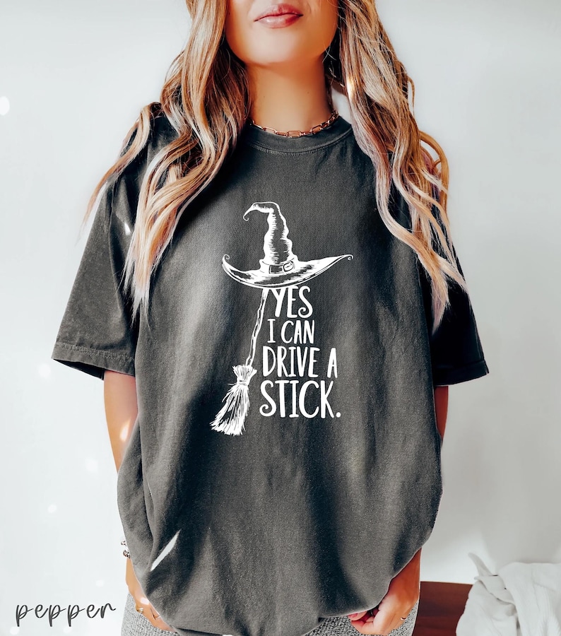 以下が含まれることがあります： 白い魔女の帽子とほうきのグラフィックと「YES I CAN DRIVE A STICK」のテキストが特徴の濃い灰色のTシャツ。 このカジュアルなTシャツは、ハロウィーンや魔女をテーマにしたイベントに最適です。