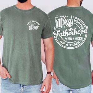May include: A sage green t-shirt with white graphic text and illustrations. The front features the word "FATHERHOOD" above a beer mug and skeleton hand. The back reads "SURVIVING FATHERHOOD ONE BEER AT A TIME" in a circular design.