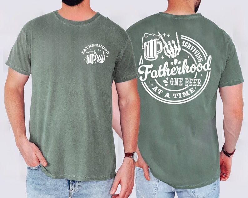 May include: A sage green t-shirt with white graphic text and illustrations. The front features the word "FATHERHOOD" above a beer mug and skeleton hand. The back reads "SURVIVING FATHERHOOD ONE BEER AT A TIME" in a circular design.