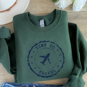 Puede incluir: Una sudadera verde oscuro con un sello desgastado azul marino que dice "Time to Travel" con un gr&aacute;fico de avi&oacute;n dentro del sello.