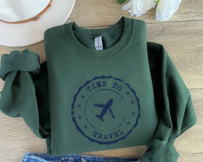 Puede incluir: Una sudadera verde oscuro con un sello desgastado azul marino que dice "Time to Travel" con un gr&aacute;fico de avi&oacute;n dentro del sello.