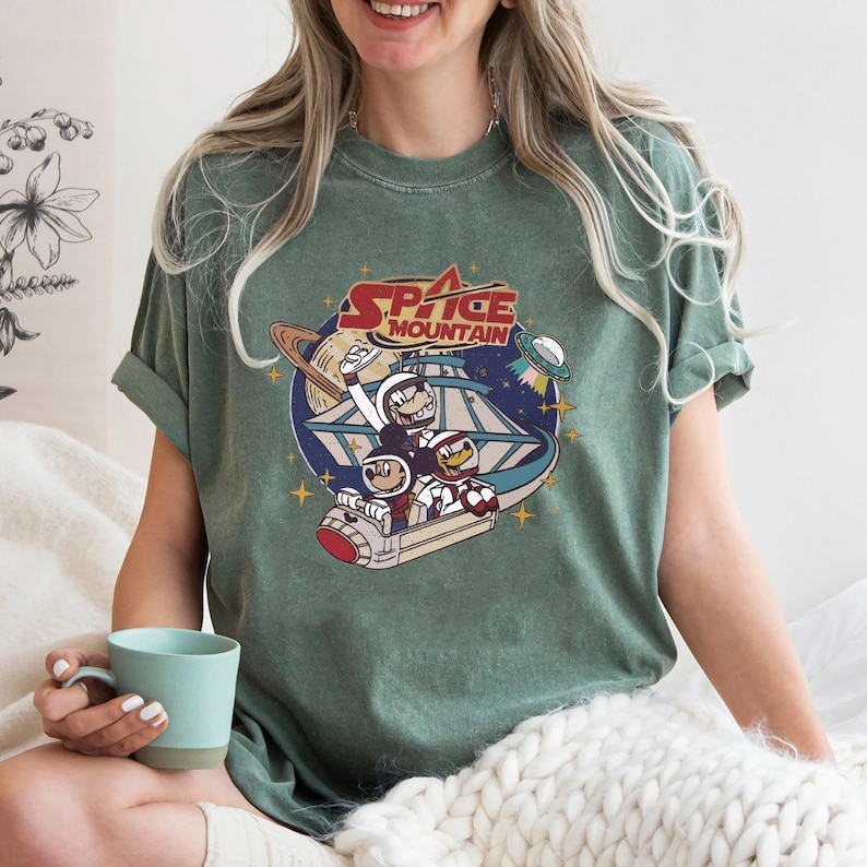 May include: A green t-shirt with a vintage-style graphic of Mickey Mouse and Donald Duck riding Space Mountain. The text "Space Mountain" is printed in red and white.