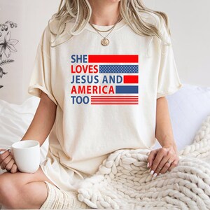 Może przedstawiać: Biały T-shirt z nadrukiem "SHE LOVES JESUS AND AMERICA TOO" w kolorze czerwonym, niebieskim i białym. Tekst jest otoczony wzorem amerykańskiej flagi w kolorze czerwonym, białym i niebieskim.