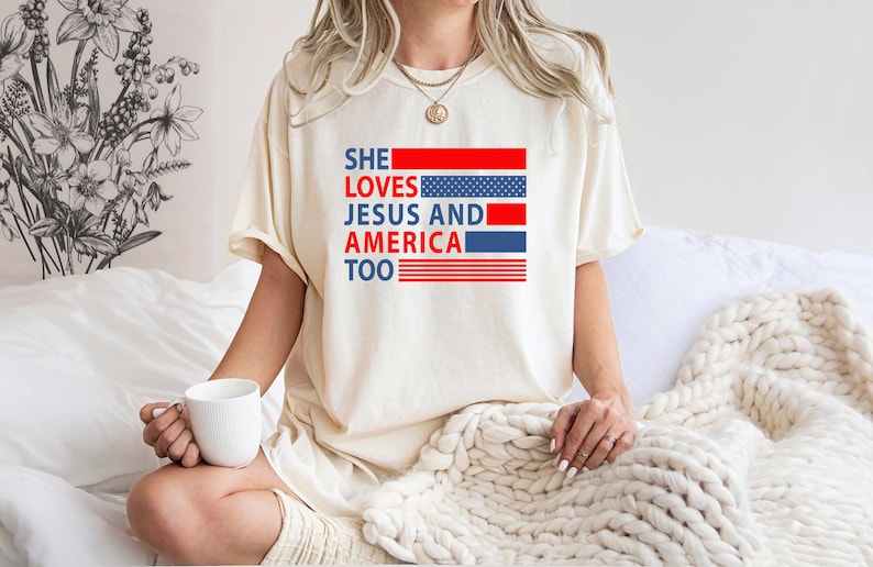 Może przedstawiać: Biały T-shirt z nadrukiem "SHE LOVES JESUS AND AMERICA TOO" w kolorze czerwonym, niebieskim i białym. Tekst jest otoczony wzorem amerykańskiej flagi w kolorze czerwonym, białym i niebieskim.