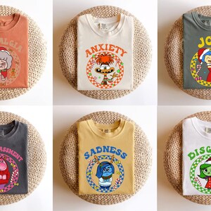 May include: Six different t-shirts with various emotions printed on them. Each shirt has a different color and features a cartoon character with a holiday theme. The emotions are Nostalgia, Anxiety, Joy, Embarrassment, Sadness, and Disgust.