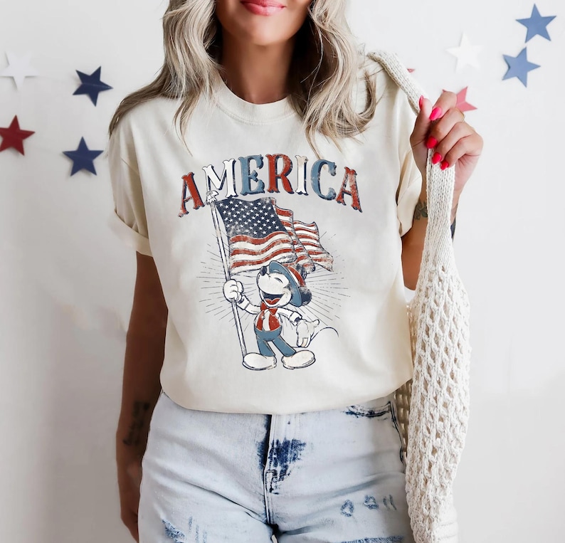 Peut inclure: T-shirt cr&egrave;me avec le mot "AMERICA" en lettres rouges et bleues. Le t-shirt pr&eacute;sente un dessin d'un personnage de dessin anim&eacute; tenant un drapeau am&eacute;ricain. Le design a un aspect vintage et vieilli.