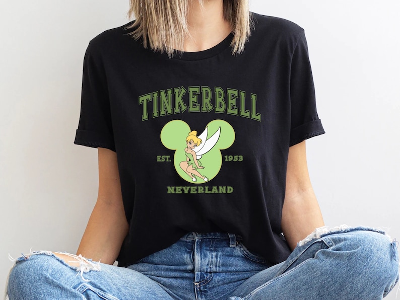 May include: Black t-shirt with a green Tinkerbell graphic inside a green Mickey Mouse ear silhouette. The text "Tinkerbell" is in green above the graphic and "EST. 1953 NEVERLAND" is below the graphic.