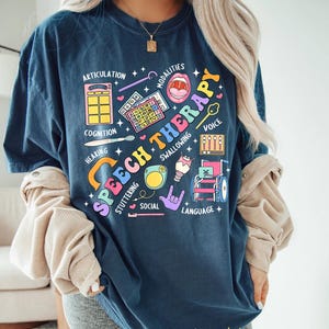 May include: A navy blue t-shirt with a colorful graphic design that says "Speech Therapy". The graphic includes illustrations of various speech therapy tools and concepts, such as a mouth, a microphone, a wheelchair, and a sign language hand gesture. The text around the graphic includes words like "articulation", "cognition", "hearing", "voice", "swallowing", "stuttering", "social", and "language".