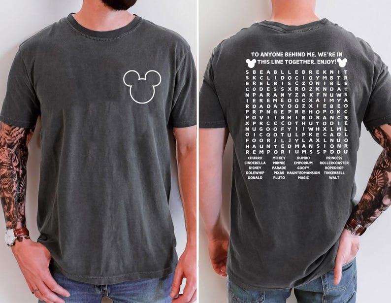 May include: Dark gray t-shirt with a white Mickey Mouse outline on the front. The back of the shirt features a word search with Disney-related words and the text "TO ANYONE BEHIND ME. WE'RE IN THIS LINE TOGETHER. ENJOY!"