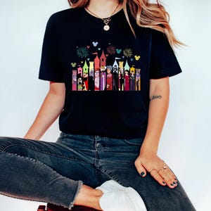 Puede incluir: Camiseta negra con un gr&aacute;fico colorido de personajes de dibujos animados y torres de castillo. El dise&ntilde;o incluye fuegos artificiales y siluetas de Mickey Mouse. La camiseta la lleva una persona sentada en un taburete rojo.