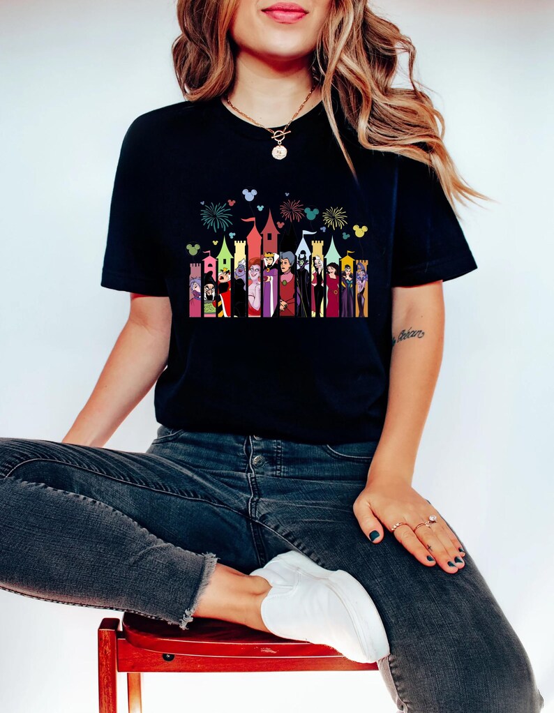 Puede incluir: Camiseta negra con un gr&aacute;fico colorido de personajes de dibujos animados y torres de castillo. El dise&ntilde;o incluye fuegos artificiales y siluetas de Mickey Mouse. La camiseta la lleva una persona sentada en un taburete rojo.