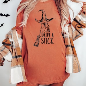 以下が含まれることがあります： 黒い魔女の帽子とほうきのグラフィックが入ったオレンジ色のTシャツ。シャツのテキストは「YES I CAN DRIVE A STICK」と書かれています。その人はTシャツの上にチェックのシャツを着ています。背景には黒いコウモリの装飾があります。
