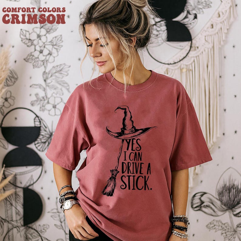 以下が含まれることがあります： 「YES I CAN DRIVE A STICK」のテキストと魔女の帽子とほうきのグラフィックが入ったクリムゾンカラーのTシャツ。シャツはComfort Colorsブランドです。