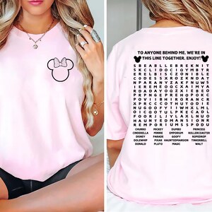 Puede incluir: Camiseta rosa claro con un contorno negro de Minnie Mouse y un lazo en la parte delantera. La parte trasera presenta una b&uacute;squeda de palabras con tem&aacute;tica de Disney y el texto "TO ANYONE BEHIND ME, WE'RE IN THIS LINE TOGETHER. ENJOY!"