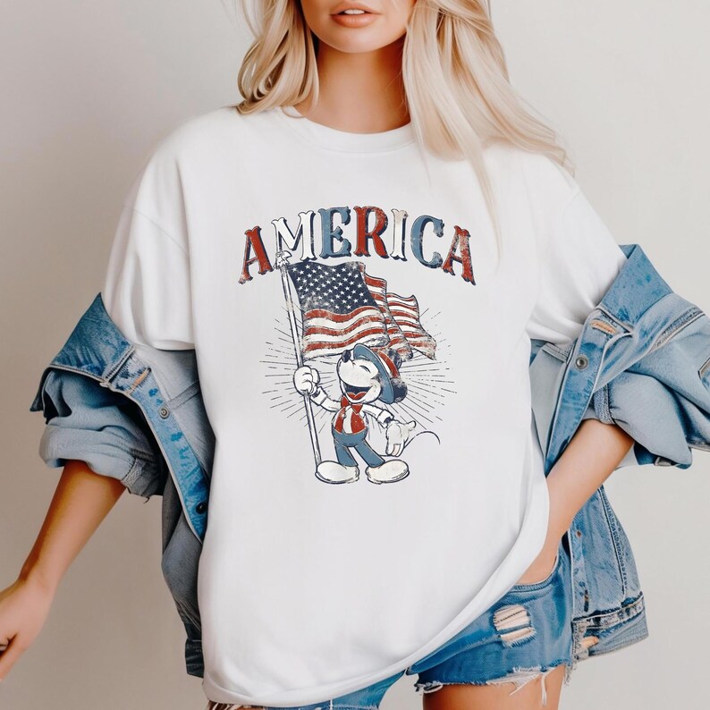 Peut inclure: T-shirt blanc avec un motif vieilli de Mickey Mouse tenant un drapeau am&eacute;ricain, avec le mot "AMERICA" au-dessus. Le design utilise des couleurs rouge, blanc et bleu, &eacute;voquant un th&egrave;me patriotique. Le t-shirt a un col rond classique et des manches courtes.