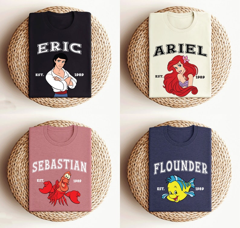 May include: Four different t-shirts with a Disney theme. The shirts are black, cream, pink, and navy blue. Each shirt has a different character from the Little Mermaid movie. The shirts have the character's name and the year 1989 printed on them.