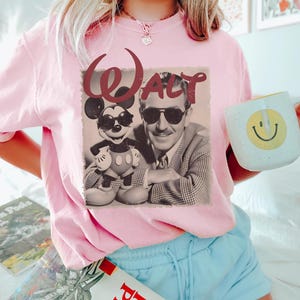 May include: A light pink t-shirt featuring a graphic print of Walt Disney and Mickey Mouse both wearing sunglasses. The word WALT is printed in a dark red, stylized font above the figures. The image is rendered in a sepia-toned, vintage style.