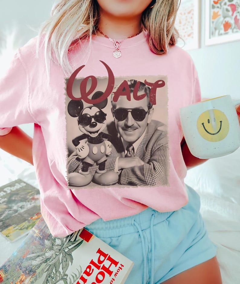 May include: A light pink t-shirt featuring a graphic print of Walt Disney and Mickey Mouse both wearing sunglasses. The word WALT is printed in a dark red, stylized font above the figures. The image is rendered in a sepia-toned, vintage style.