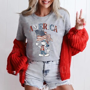 Peut inclure: Un t-shirt gris avec le mot "AMERICA" en lettres rouges et bleues. Le t-shirt pr&eacute;sente un personnage de dessin anim&eacute; tenant un drapeau am&eacute;ricain. Un pull rouge est drap&eacute; sur les &eacute;paules.