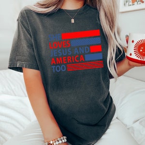 Może przedstawiać: Szary t-shirt z nadrukiem "She loves Jesus and America too" w kolorze czerwonym i niebieskim. Tekst jest ułożony w pionowe pasy z czerwonymi i niebieskimi paskami za tekstem.