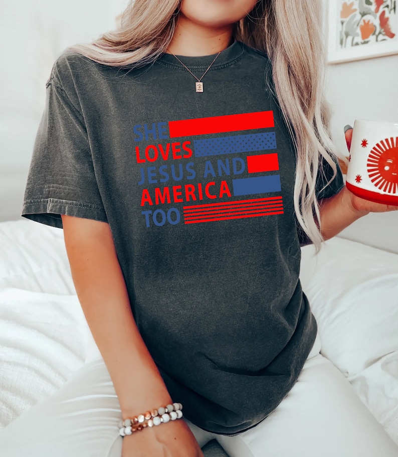Może przedstawiać: Szary t-shirt z nadrukiem "She loves Jesus and America too" w kolorze czerwonym i niebieskim. Tekst jest ułożony w pionowe pasy z czerwonymi i niebieskimi paskami za tekstem.