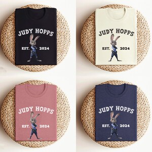May include: Four t-shirts with different colors, black, beige, pink, and navy blue, featuring a cartoon image of Judy Hopps from the movie Zootopia. The text on the shirts reads "Judy Hopps" with "Est. 2024" underneath.