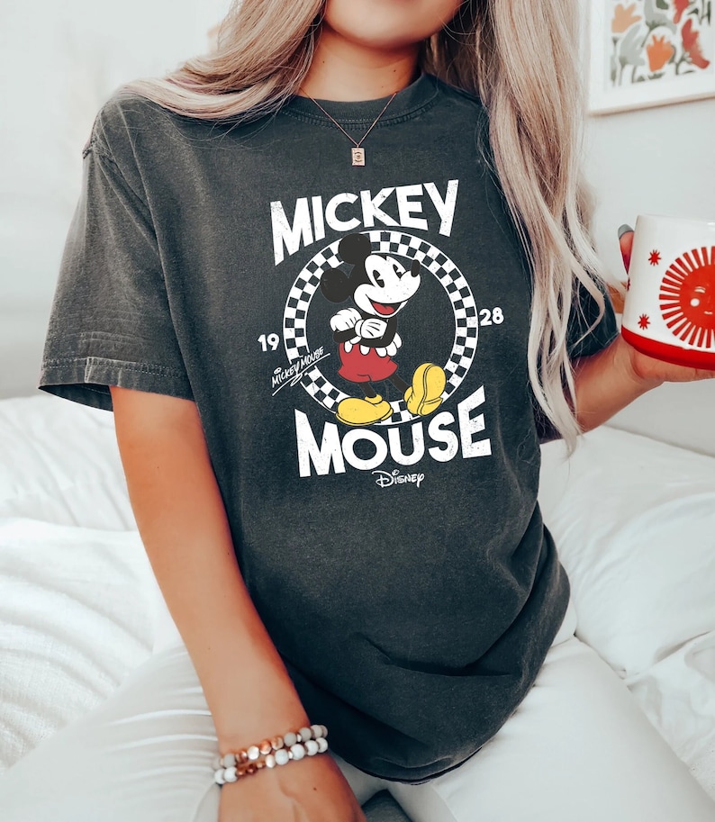 K&ouml;nnte beinhalten: Ein schwarzes T-Shirt mit einem Vintage-Mickey-Mouse-Motiv. Das Motiv zeigt Mickey Mouse in einem karierten Kreis mit dem Text "Mickey Mouse" und "Disney" darunter.