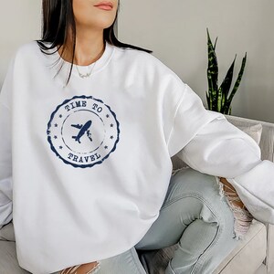 Puede incluir: Sudadera blanca con un dise&ntilde;o de sello azul desgastado que dice "Time to Travel" con un avi&oacute;n dentro del sello.