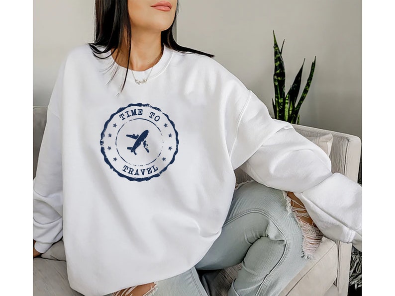 Puede incluir: Sudadera blanca con un dise&ntilde;o de sello azul desgastado que dice "Time to Travel" con un avi&oacute;n dentro del sello.