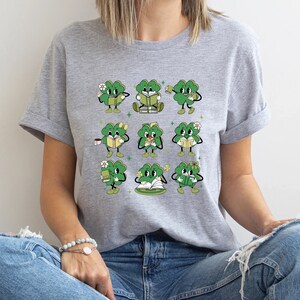 May include: A gray t-shirt with a graphic of nine green four-leaf clovers holding books, coffee cups, and flowers. The clovers have faces and are in different poses.