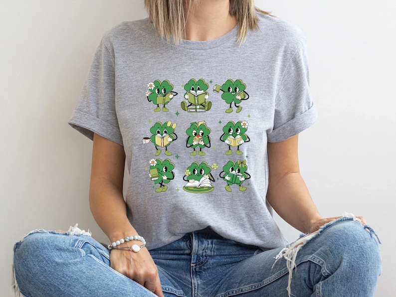 May include: A gray t-shirt with a graphic of nine green four-leaf clovers holding books, coffee cups, and flowers. The clovers have faces and are in different poses.