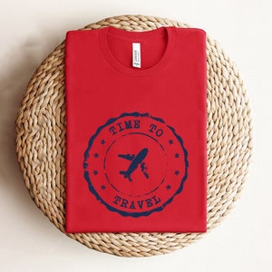 Puede incluir: Camiseta roja con un gr&aacute;fico azul y blanco de un avi&oacute;n con el texto "Time to Travel" en un c&iacute;rculo.