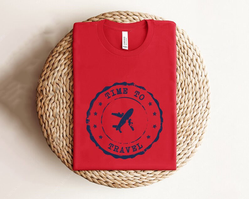 Puede incluir: Camiseta roja con un gr&aacute;fico azul y blanco de un avi&oacute;n con el texto "Time to Travel" en un c&iacute;rculo.