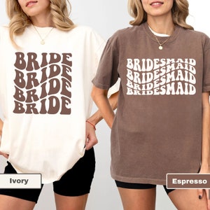 May include: Two t-shirts with a retro font design. The white t-shirt says "BRIDE" in brown text. The brown t-shirt says "BRIDESMAID" in white text. The t-shirts are shown on models wearing black shorts. The white t-shirt is labeled "Ivory" and the brown t-shirt is labeled "Espresso".