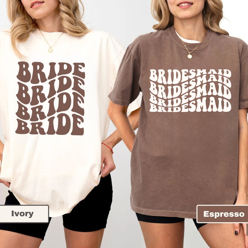 May include: Two t-shirts with a retro font design. The white t-shirt says "BRIDE" in brown text. The brown t-shirt says "BRIDESMAID" in white text. The t-shirts are shown on models wearing black shorts. The white t-shirt is labeled "Ivory" and the brown t-shirt is labeled "Espresso".