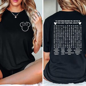 Puede incluir: Camiseta negra con un contorno blanco de Minnie Mouse y un lazo en la parte delantera. La parte trasera muestra el texto "TO ANYONE BEHIND ME. WE'RE IN THIS LINE TOGETHER. ENJOY!" y una b&uacute;squeda de palabras de Disney.