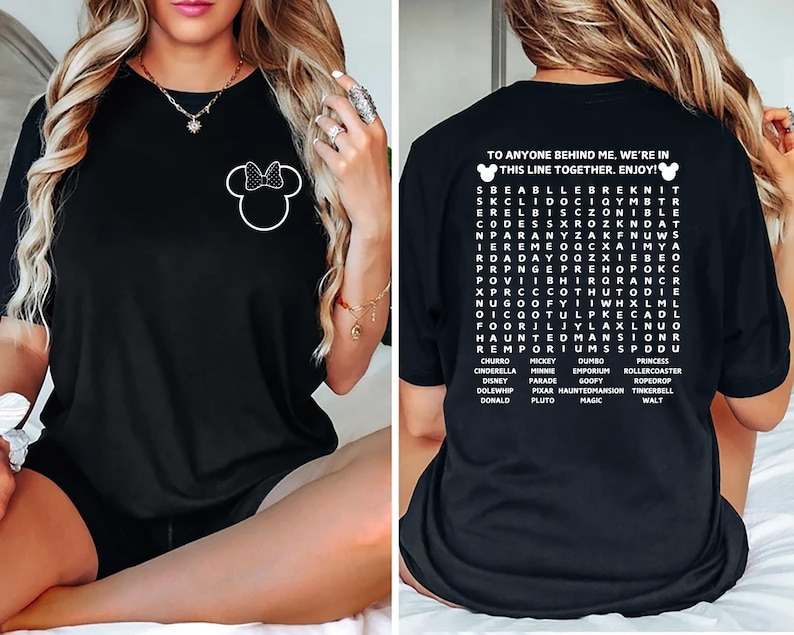 Puede incluir: Camiseta negra con un contorno blanco de Minnie Mouse y un lazo en la parte delantera. La parte trasera muestra el texto "TO ANYONE BEHIND ME. WE'RE IN THIS LINE TOGETHER. ENJOY!" y una b&uacute;squeda de palabras de Disney.