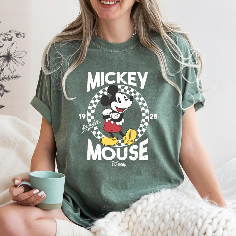K&ouml;nnte beinhalten: Ein gr&uuml;nes T-Shirt mit einem Vintage-Grafikdruck von Mickey Mouse. Die Grafik zeigt Mickey Mouse in einem karierten Kreis mit dem Text "Mickey Mouse" und "Disney" darunter.