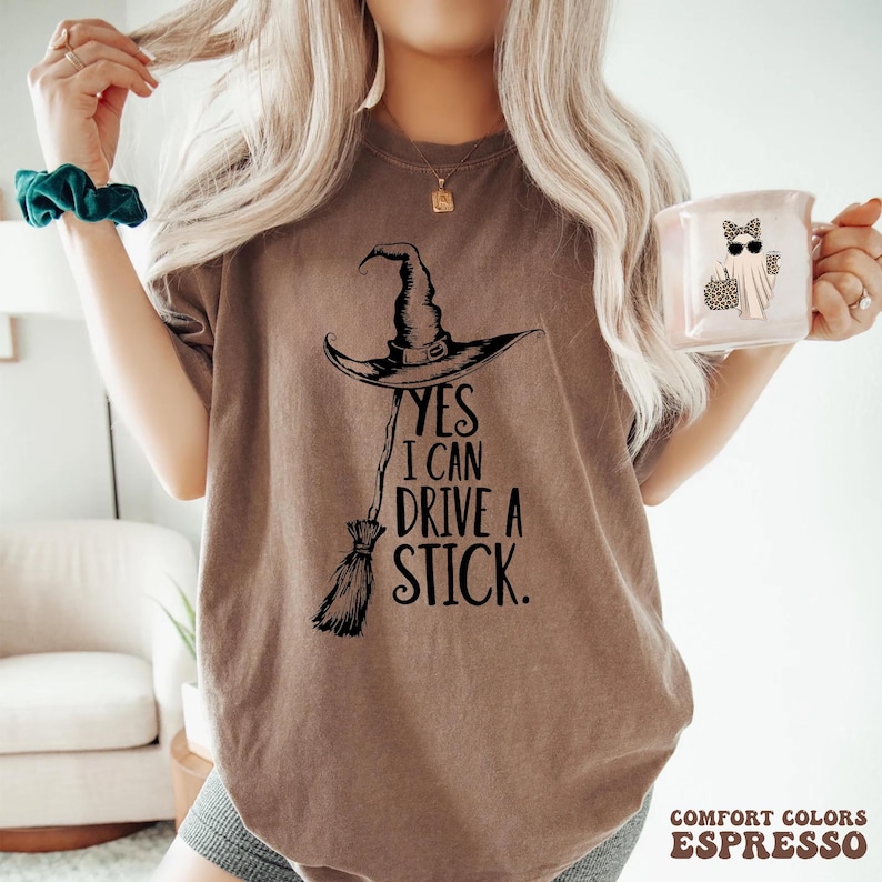 以下が含まれることがあります： エスプレッソ色のTシャツに、黒い魔女の帽子とほうきのグラフィック。テキストは「YES I CAN DRIVE A STICK」と書かれています。サングラスとヒョウ柄のバッグを身に着けた幽霊が描かれた白いマグカップが手に持たれています。