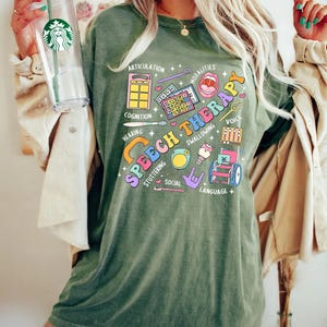 May include: Green t-shirt with a colorful graphic design that says "Speech Therapy" and includes icons representing different aspects of speech therapy, such as articulation, cognition, hearing, swallowing, stuttering, social, language, and voice.