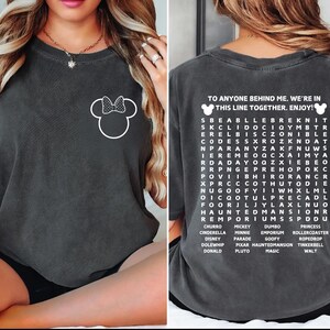 Puede incluir: Camiseta gris oscuro con un contorno blanco de Minnie Mouse en la parte delantera. La parte posterior de la camiseta tiene un juego de b&uacute;squeda de palabras con palabras tem&aacute;ticas de Disney y el texto "TO ANYONE BEHIND ME. WE'RE IN THIS LINE TOGETHER. ENJOY!"