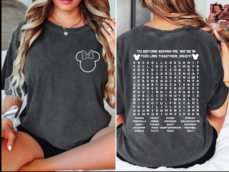 Puede incluir: Camiseta gris oscuro con un contorno blanco de Minnie Mouse en la parte delantera. La parte posterior de la camiseta tiene un juego de b&uacute;squeda de palabras con palabras tem&aacute;ticas de Disney y el texto "TO ANYONE BEHIND ME. WE'RE IN THIS LINE TOGETHER. ENJOY!"